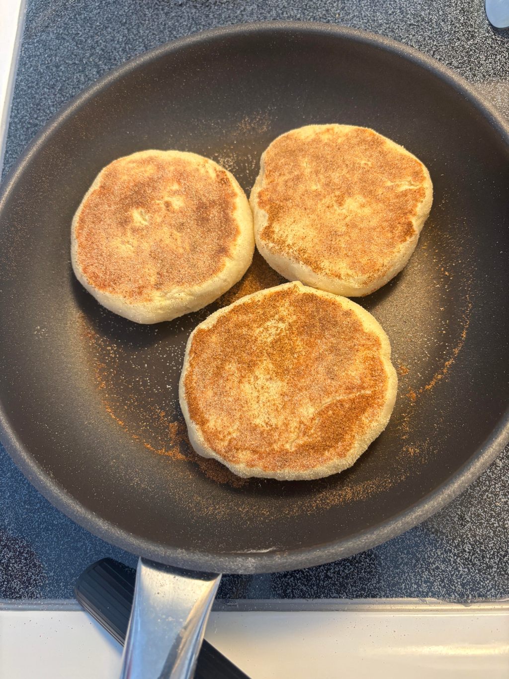Three homemade English muffins dusted with cornmeal, browning in a skillet